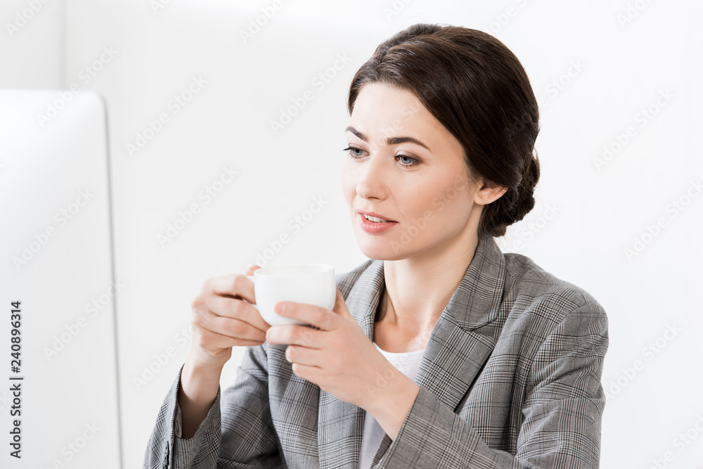 attractive businesswoman in grey suit sitting at table with computer and holding cup of coffee in office attractive businesswoman in grey suit sitting at table with computer and holding cup of coffee in office
