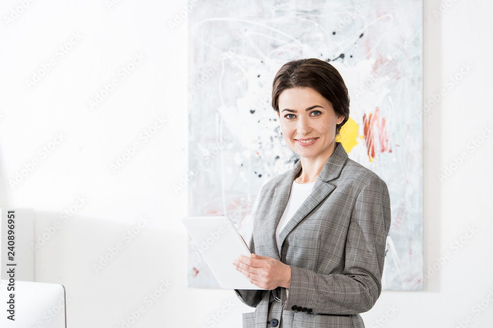 cheerful attractive businesswoman in grey suit holding tablet and looking at camera in office cheerful attractive businesswoman in grey suit holding tablet and looking at camera in office