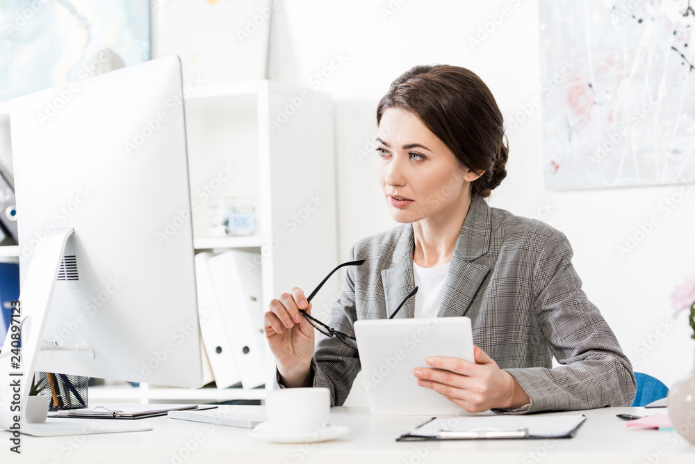 beautiful businesswoman in grey suit holding tablet and looking at computer in office beautiful businesswoman in grey suit holding tablet and looking at computer in office