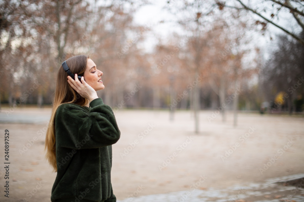 Side view of pretty young female in headphones smiling and listening to music while standing on blurred background of autumn park Side view of pretty young female in headphones smiling and listening to music while standing on blurred background of autumn park