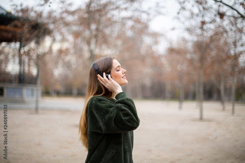 Side view of pretty young female in headphones smiling and listening to music while standing on blurred background of autumn park Side view of pretty young female in headphones smiling and listening to music while standing on blurred background of autumn park