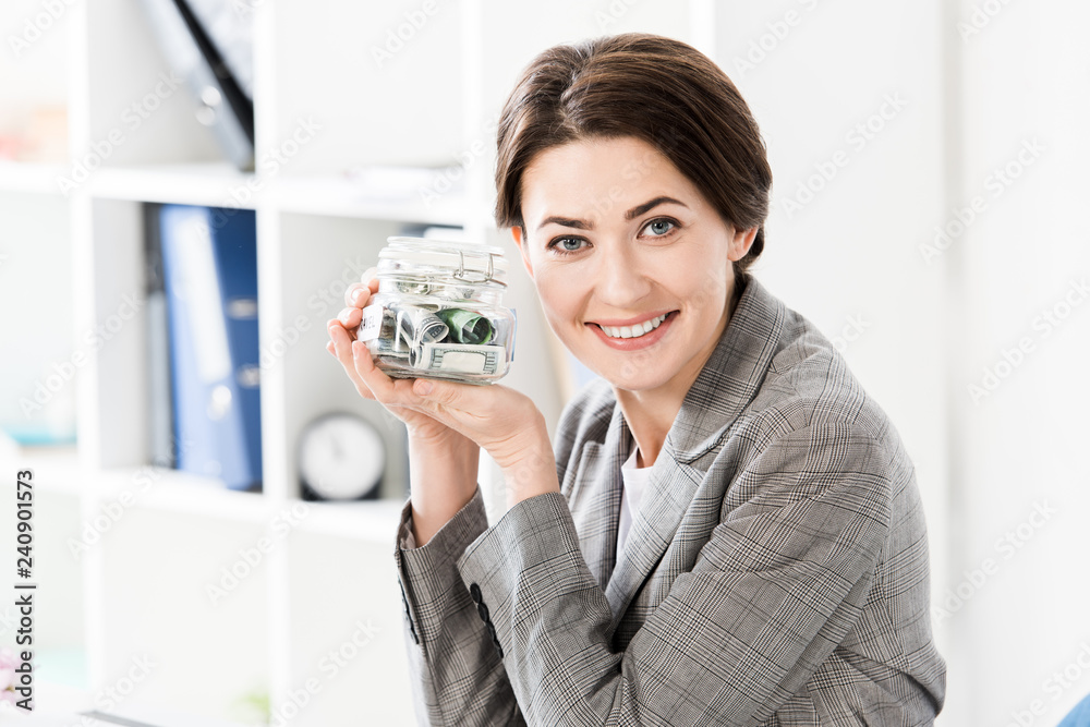 attractive happy businesswoman holding glass jar with money in office and looking at camera attractive happy businesswoman holding glass jar with money in office and looking at camera