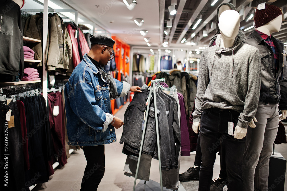 Stylish casual african american man at jeans jacket and black beret at clothes store looking on new jacket against mannequin.