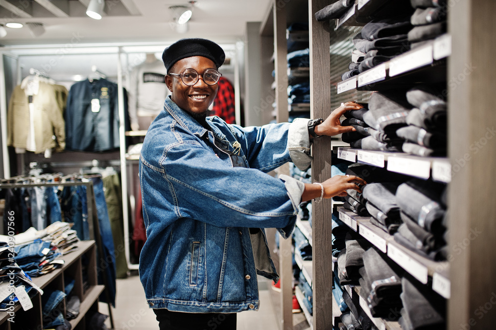 Stylish casual african american man at jeans jacket and black beret at clothes store looking on pants at shelves.