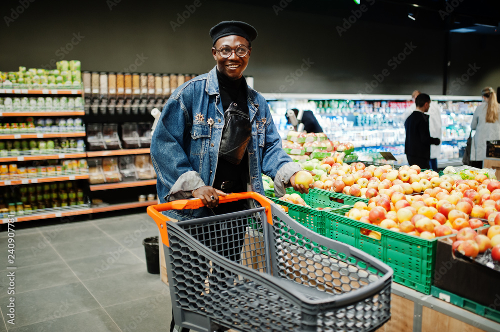 Stylish casual african american man at jeans jacket and black beret at organic section of supermarket with shopping trolley cart.