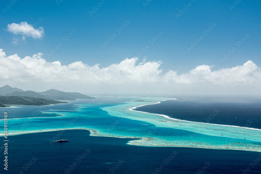 Aerial view on lagoon of Raiatea island in French Polynesia with blue and turquoise water, barrier reef, blue sky, hills with tropical forest and white clouds