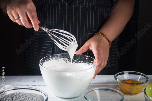Fényképezés Female chef whipping egg whites  to soft peaks in glass bowl with whisk and hand on white wooden table