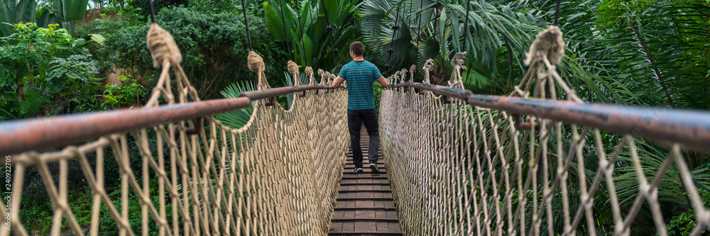 Naklejka premium Młody człowiek przechodzi przez wiszący most w panoramie dżungli