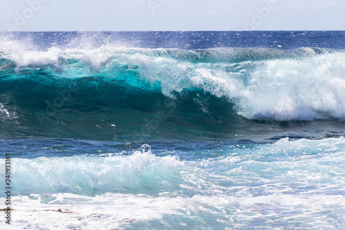Wave breaking near shore on South Point, on Hawaii's Big Island. Sea foam in foreground. Foam on top of wave's clear blue-green water; deep blue Pacific ocean in the background.  