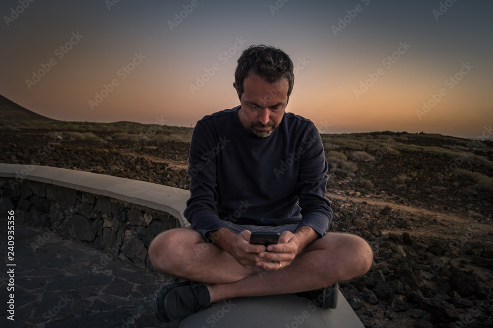 Man sitting legs crossed using smartphone with a sunset on the ocean in background. Caucasian happy boy enjoying free time outdoor on a summer evening near the sea, writing messages with cell phone.