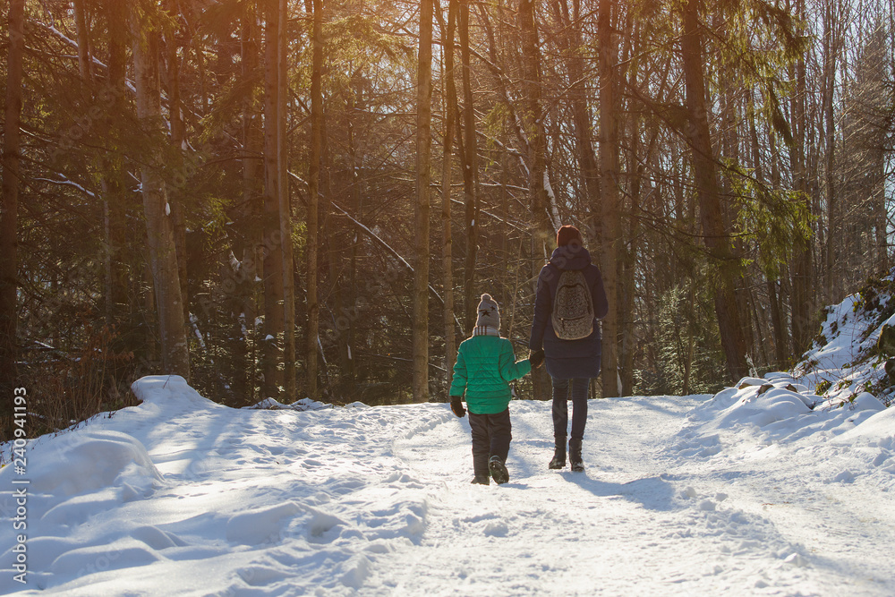 Mother with son walking along snow-covered road against the background of coniferous forest. Winter sunny day
