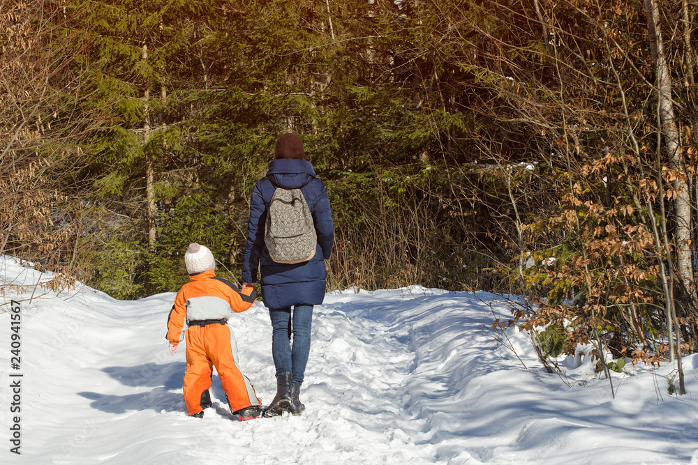 Mother withlittle son standing along snow-covered road against the background of coniferous forest. Winter sunny day