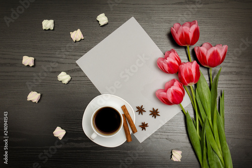 Holiday concept. Bouquet of pink tulips, a cup of coffee, marshmallow and sheet of paper on a black wooden background. Directly above.