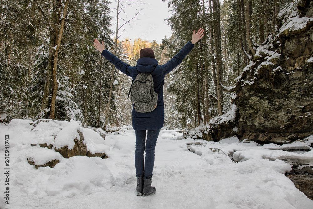 Girl with a backpack is standing with arms raised in a snowy forest. Winter day