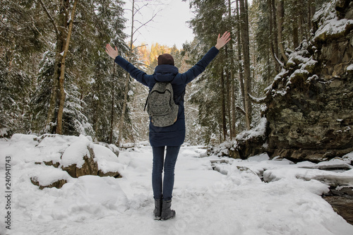 Girl with a backpack is standing with arms raised in a snowy forest. Winter day