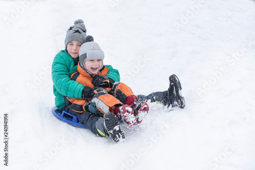 Two joyful boys slides down the hill on snow saucer. Brotherly friendship. Winter day