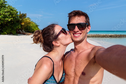 happy young couple taking a selfie, tropical island and clear blue water as background. Girl kissing his boyfriend