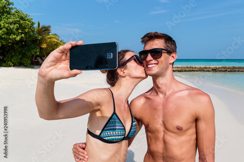 happy young couple taking a selfie, tropical island and clear blue water as background. Girl kissing his boyfriend
