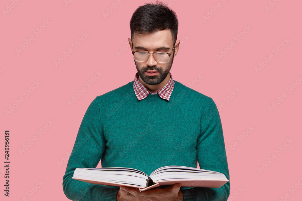 Waist up shot of serious unshaven young man gets knowledge from scientific book, wears spectacles for good vision, being diligent pupil, focused in thick textbook, isolated over pink background Waist up shot of serious unshaven young man gets knowledge from scientific book, wears spectacles for good vision, being diligent pupil, focused in thick textbook, isolated over pink background