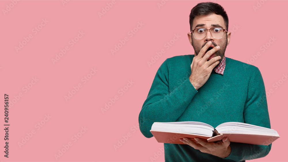 Photo of surprised European man covers mouth with palm, has scared expression, gazes at camera with eyes full of shock and fear, reads some surprised indormation in textbook, isolated on pink