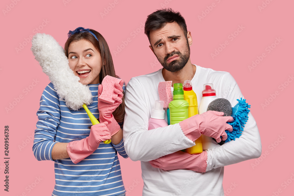 Positive smiling woman carries white brush, rejoices good results after cleaning, upset bearded guy carries detergents, looks fatigue, isolated over pink background. People, team work, cleaning