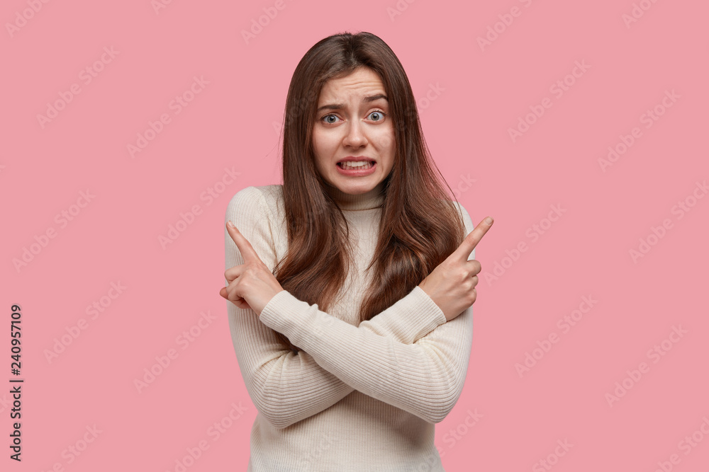 Puzzled lady crosses hands over chest, points in different sides left and right, hesitates while makes important choice, clenches teeth nervously, isolated on pink background. Woman gives directions