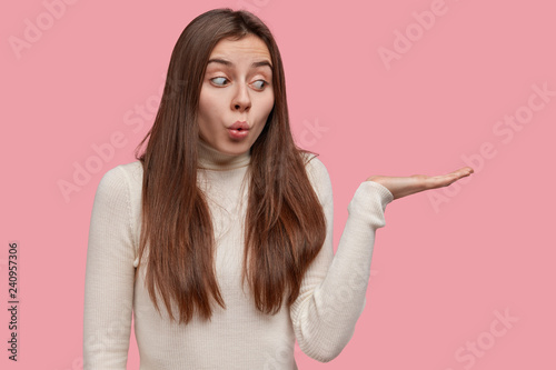 Shocked attractive woman has surprised expression, holds copy space, raises palm, pretends holding something, has appealing look, isolated on pink background with copy space for your promotion