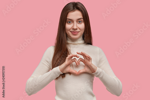 Isolated shot of friendly looking young girl shows heart gesture over chest, ...