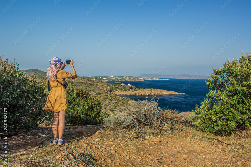 travel girl in yellow dress back to camera taking photo on mobile device of beautiful picturesque south European Mediterranean sea scenic landscape shoreline from above top point of mountain cape