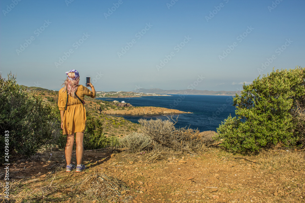 travel girl in yellow dress back to camera taking photo on mobile device of beautiful picturesque south European Mediterranean sea scenic landscape shoreline from above top point of mountain cape