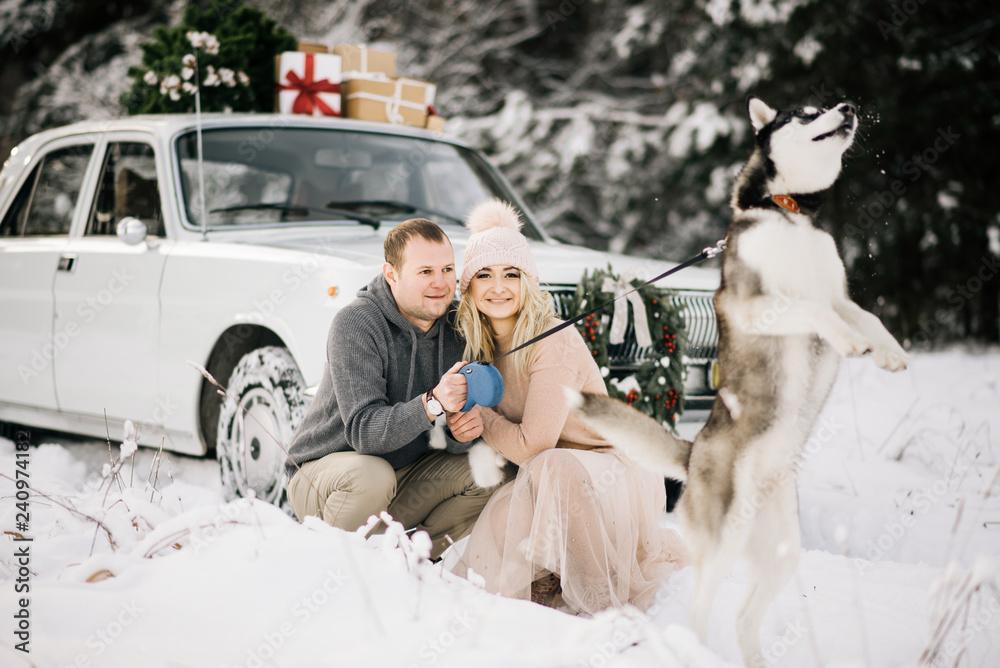 A guy and a girl getting ready for Christmas, walking the dog husky on a background of vintage car, on the roof tree and gifts in the winter snowy forest
