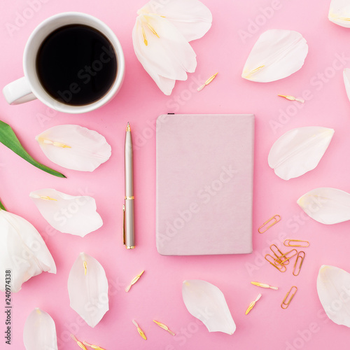 White flowers and petals with mug of coffee, notebook and pen on pink background. Flat lay, top view.