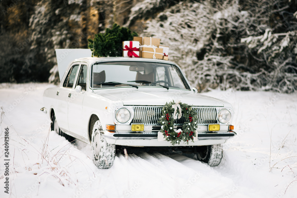 Retro car with gifts and Christmas tree in winter snowy forest and beautiful Christmas wreath. Holiday decor, Santa Claus delivery Retro car with gifts and Christmas tree in winter snowy forest and beautiful Christmas wreath. Holiday decor, Santa Claus delivery