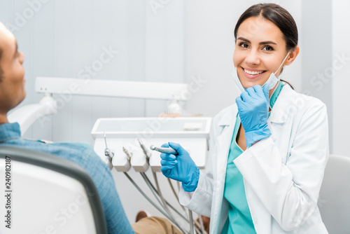 cheerful female dentist holding drill and smiling near patient photo