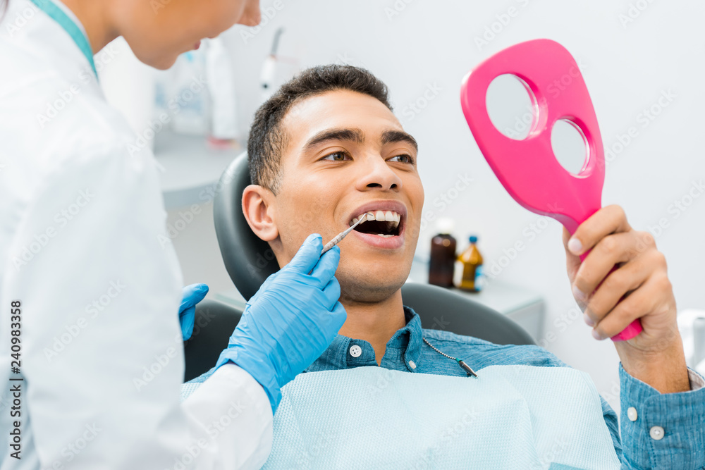 © LIGHTFIELD STUDIOS - female dentist holding medical instrument near african american patient looking at mirror