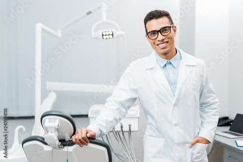 handsome african american dentist smiling and standing with hand in pocket
