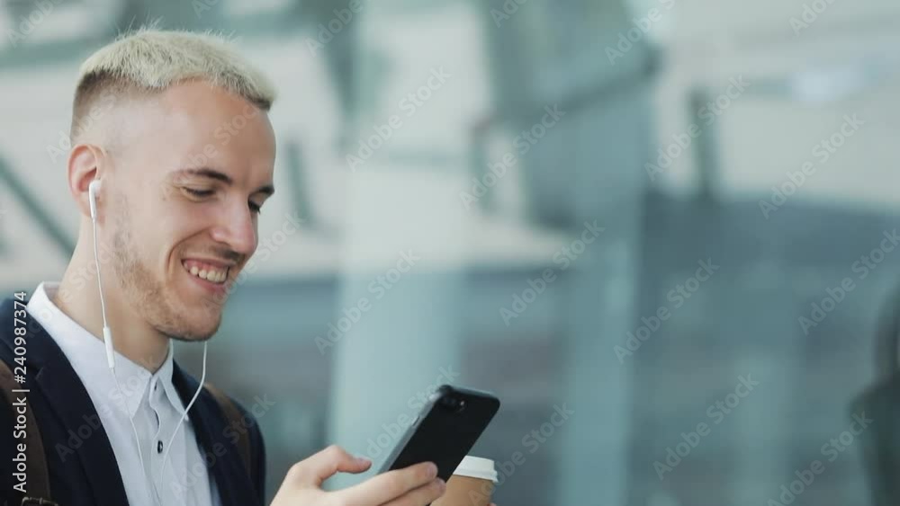Young attractive man walks down the street holding coffee and using phone. He listens to music at headphones walking around the business center Young attractive man walks down the street holding coffee and using phone. He listens to music at headphones walking around the business center