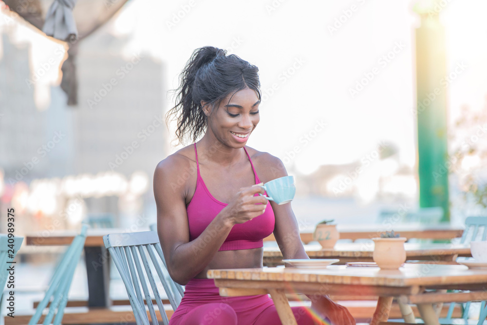 Beautiful tall athletic African American Woman wearing a bright pink  workout outfit sits in an outdoor cafe drinking coffee or tea in deep  though on a bright sunny day Photos