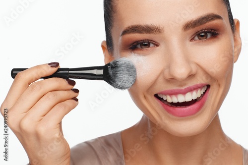Portrait of a beautiful smiling young woman holding a brush in her hand and applying powder to her face with a brush