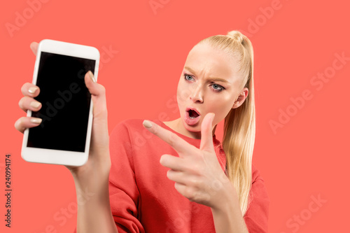 Portrait of a surprised, smiling, happy, astonished girl showing blank screen mobile phone isolated over coral background.