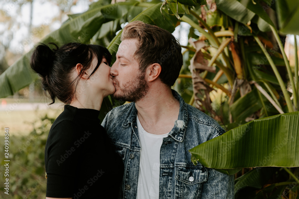 Portrait of Two Cute Modern Caucasian Beautiful Young Adult Guy Boyfriend Lady Girlfriend Couple Hugging and Kissing in Love in Nature with Green Plants Trees in the Outdoor Park Portrait of Two Cute Modern Caucasian Beautiful Young Adult Guy Boyfriend Lady Girlfriend Couple Hugging and Kissing in Love in Nature with Green Plants Trees in the Outdoor Park
