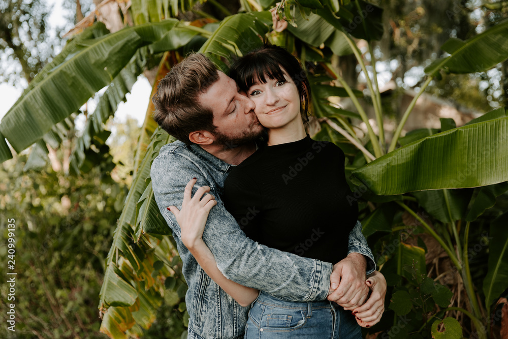 Obraz premium Portrait of Two Cute Modern Caucasian Beautiful Young Adult Guy Boyfriend Lady Girlfriend Couple Hugging and Kissing in Love in Nature with Green Plants Trees in the Outdoor Park