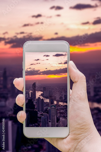 Mockup image of hand holding white mobile phone to taking a photo twilight and cityscape on highest rooftop building at Bangkok in Thailand.