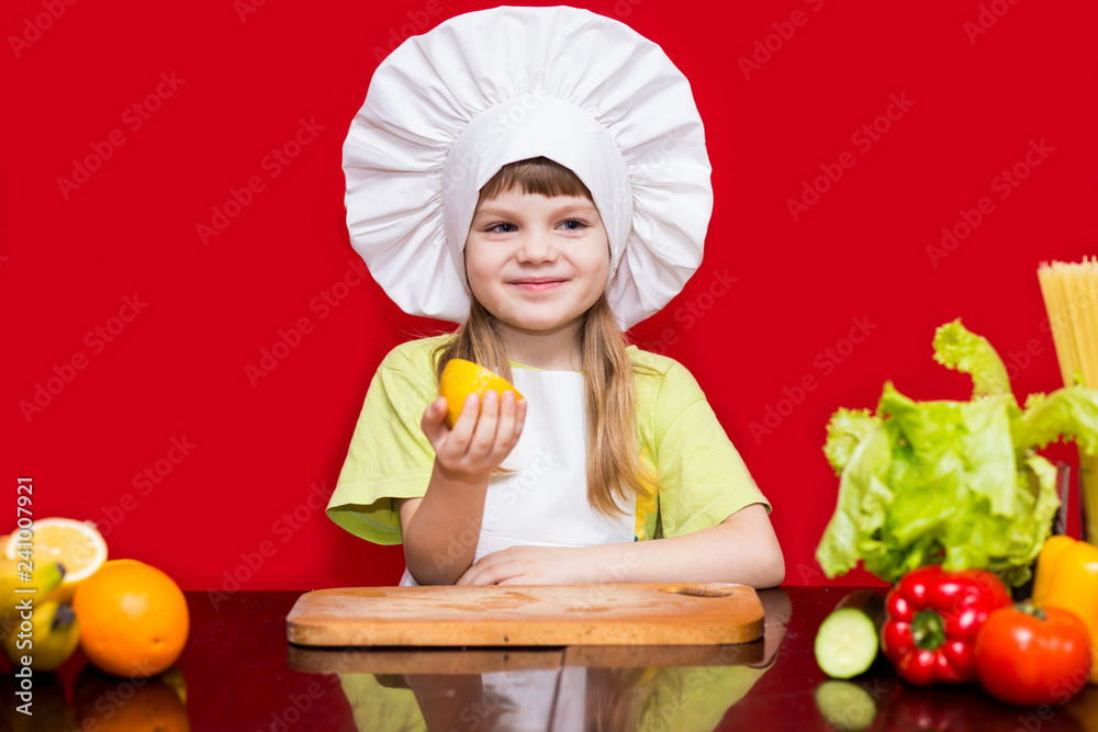 Happy little girl in chef uniform cuts fruit in kitchen. Kid chef. Cooking Concept Happy little girl in chef uniform cuts fruit in kitchen. Kid chef. Cooking Concept