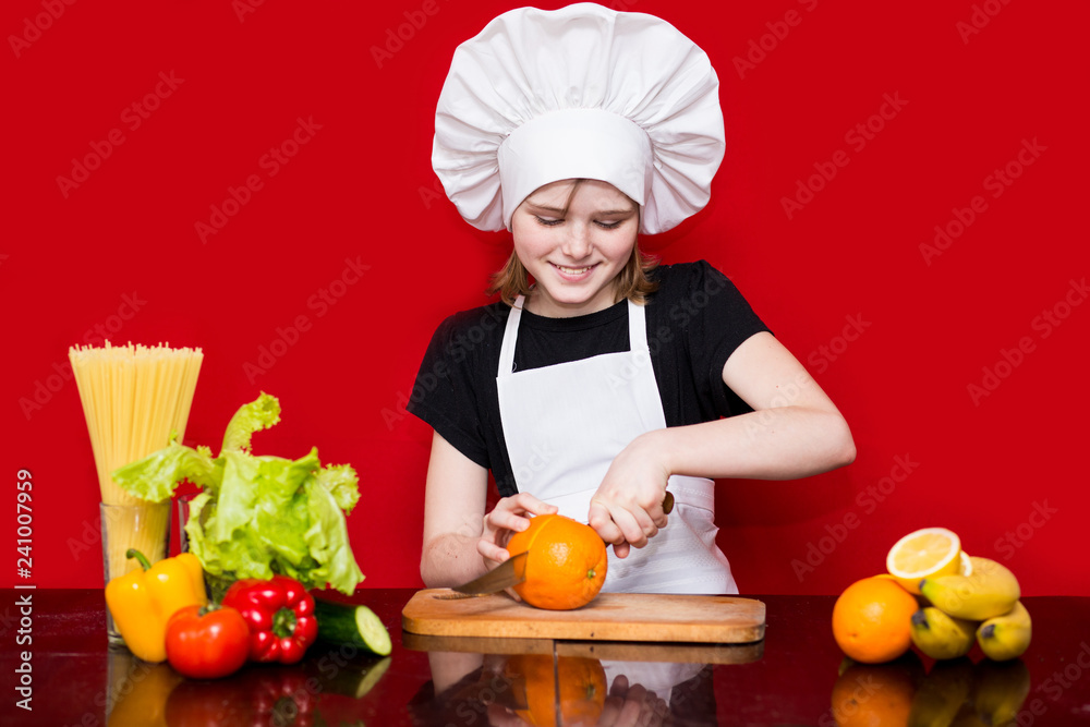 Happy little girl in chef uniform cuts fruit in kitchen. Kid chef. Cooking Concept Happy little girl in chef uniform cuts fruit in kitchen. Kid chef. Cooking Concept