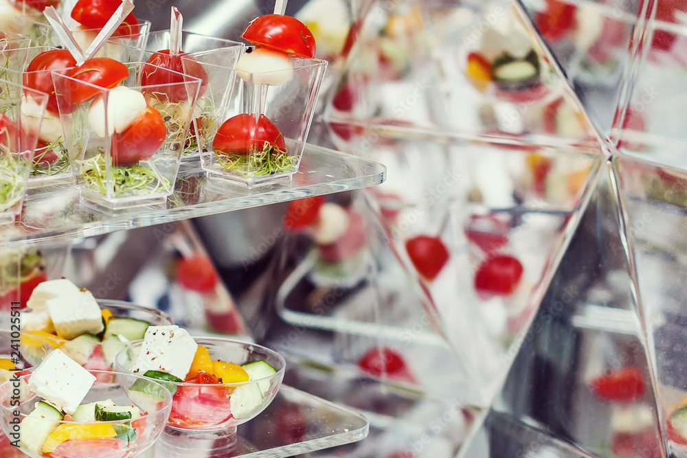 Catering for party. Close up of appetizers with cherry tomatoes, green olives, olive oil, cheese and spices in short glasses on wood brown table. Horizontal color image.