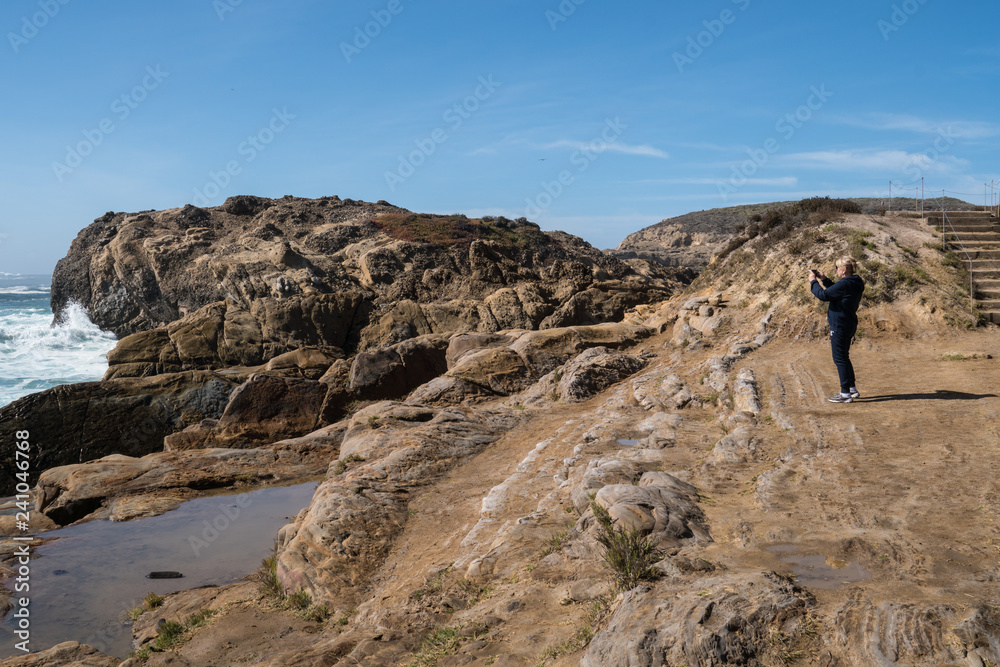 Active senior woman stands on the slippery rocks of Point Lobos State Reserve in California, admiring the scenery and taking photos with a smartphone