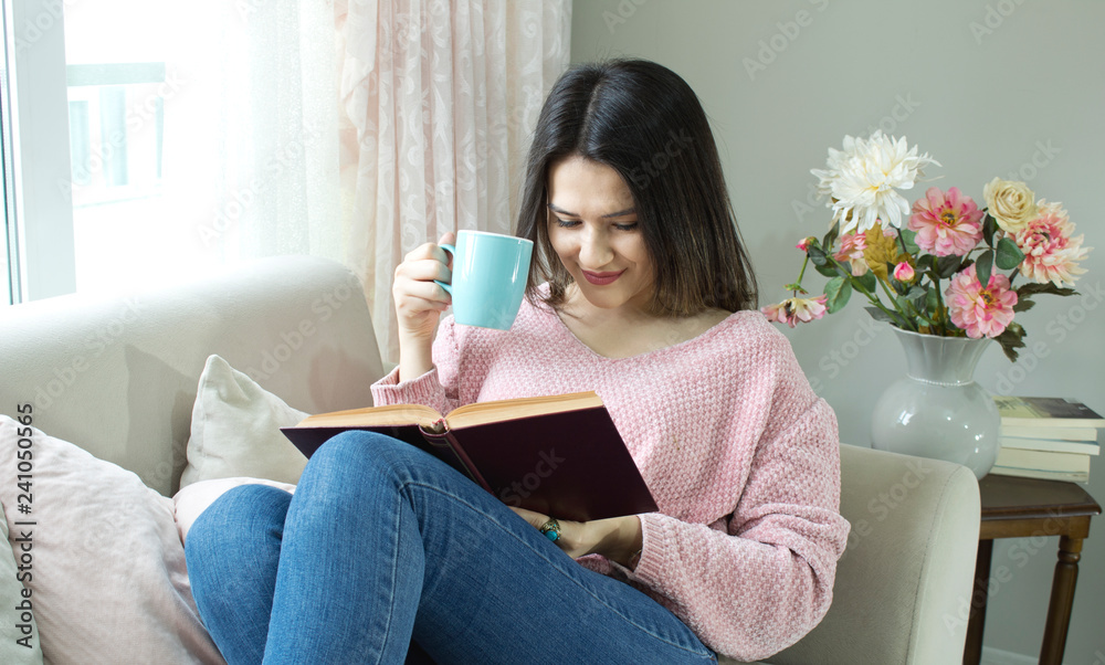 Young beautiful woman is reading book on couch and drinking coffee.