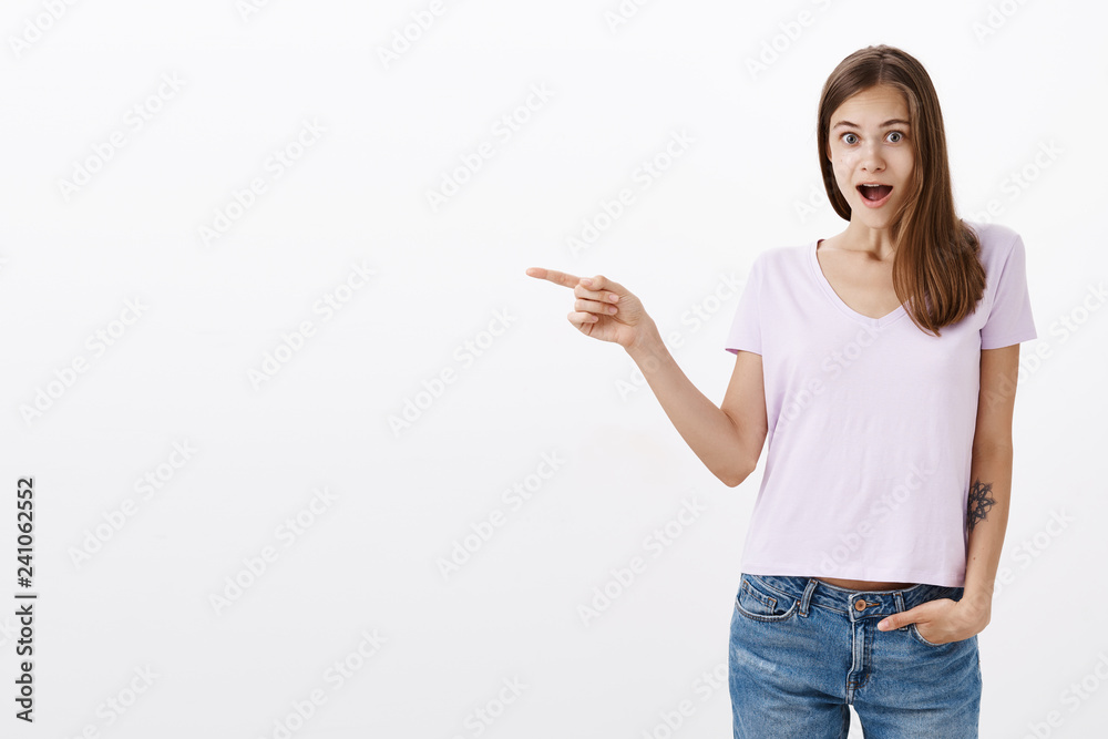 Indoor shot of surprised and amazed delighted good-looking young woman with cool tattoo on arm dropping jaw feeling amused and excited pointing left at awesome copy space against grey wall Indoor shot of surprised and amazed delighted good-looking young woman with cool tattoo on arm dropping jaw feeling amused and excited pointing left at awesome copy space against grey wall
