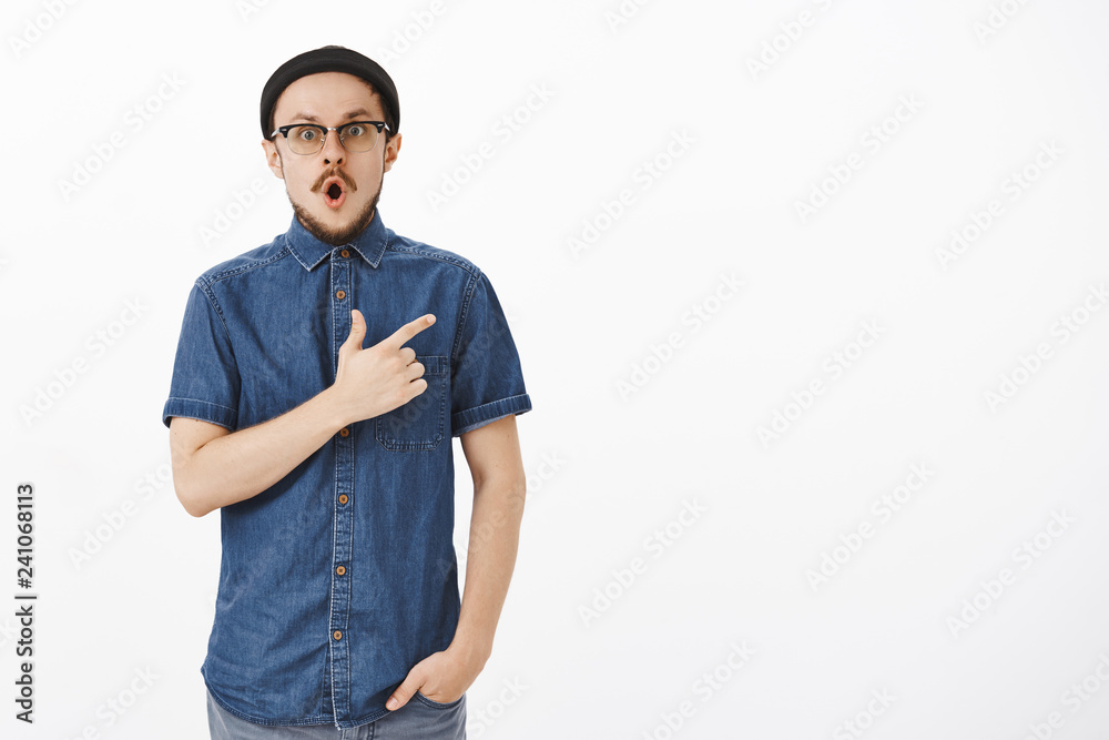 Fototapeta premium Portrait of amazed and impressed handsome young bearded guy in stylish beanie and blue shirt dropping jaw in wow sound pointing at upper right corner staring questioned and thrilled over white wall